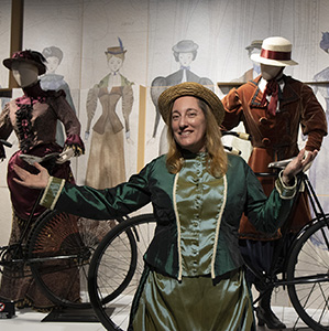 A woman in an Edwardian style costume standing in front of two costumed mannequins holding bicycles