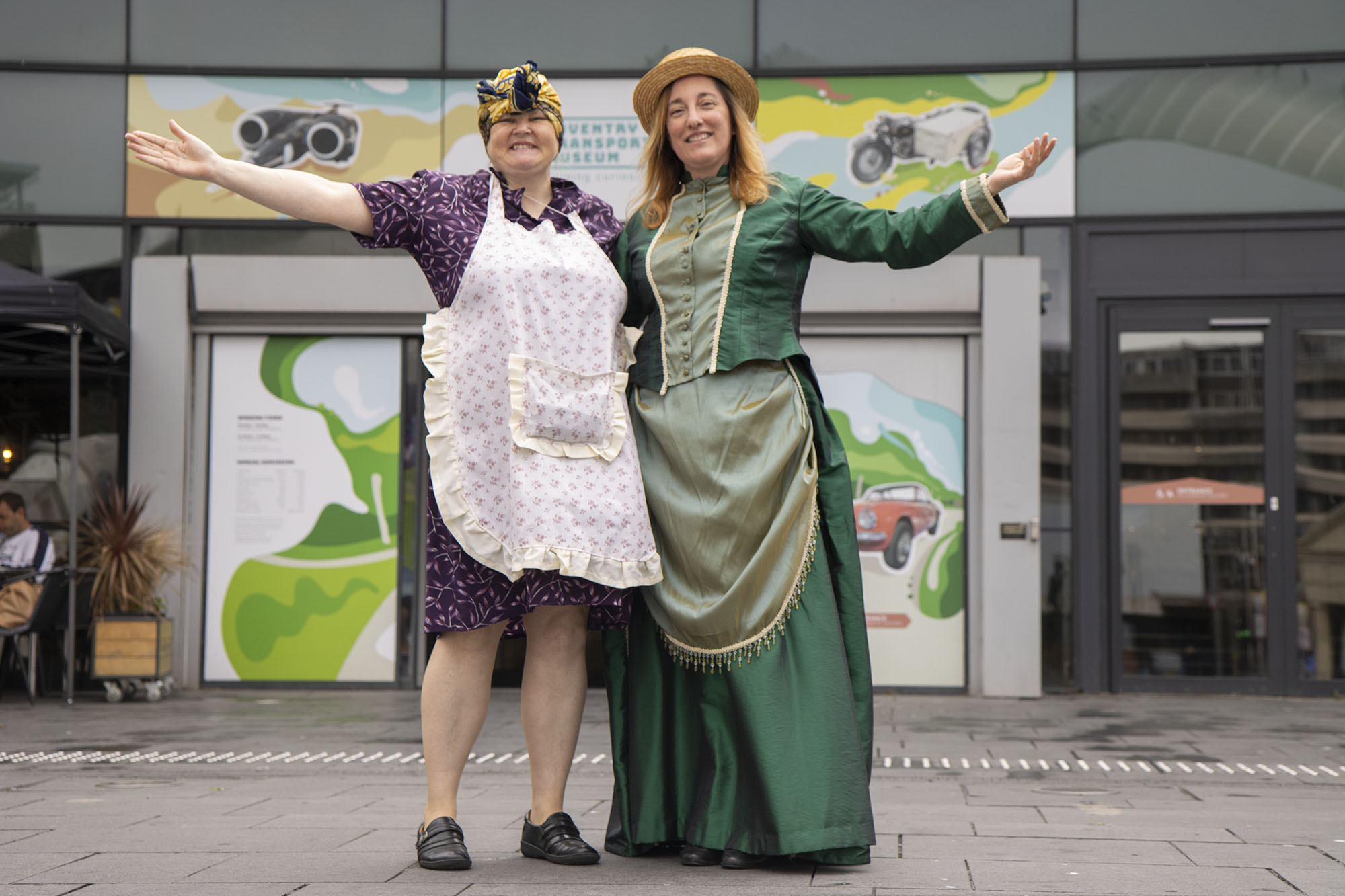 Two tour guides in costume standing outside Coventry Transport Museum with arms outstretched. The woman on the left is dressed in wartime attire, and the woman on the right is dressed as an Edwardian.