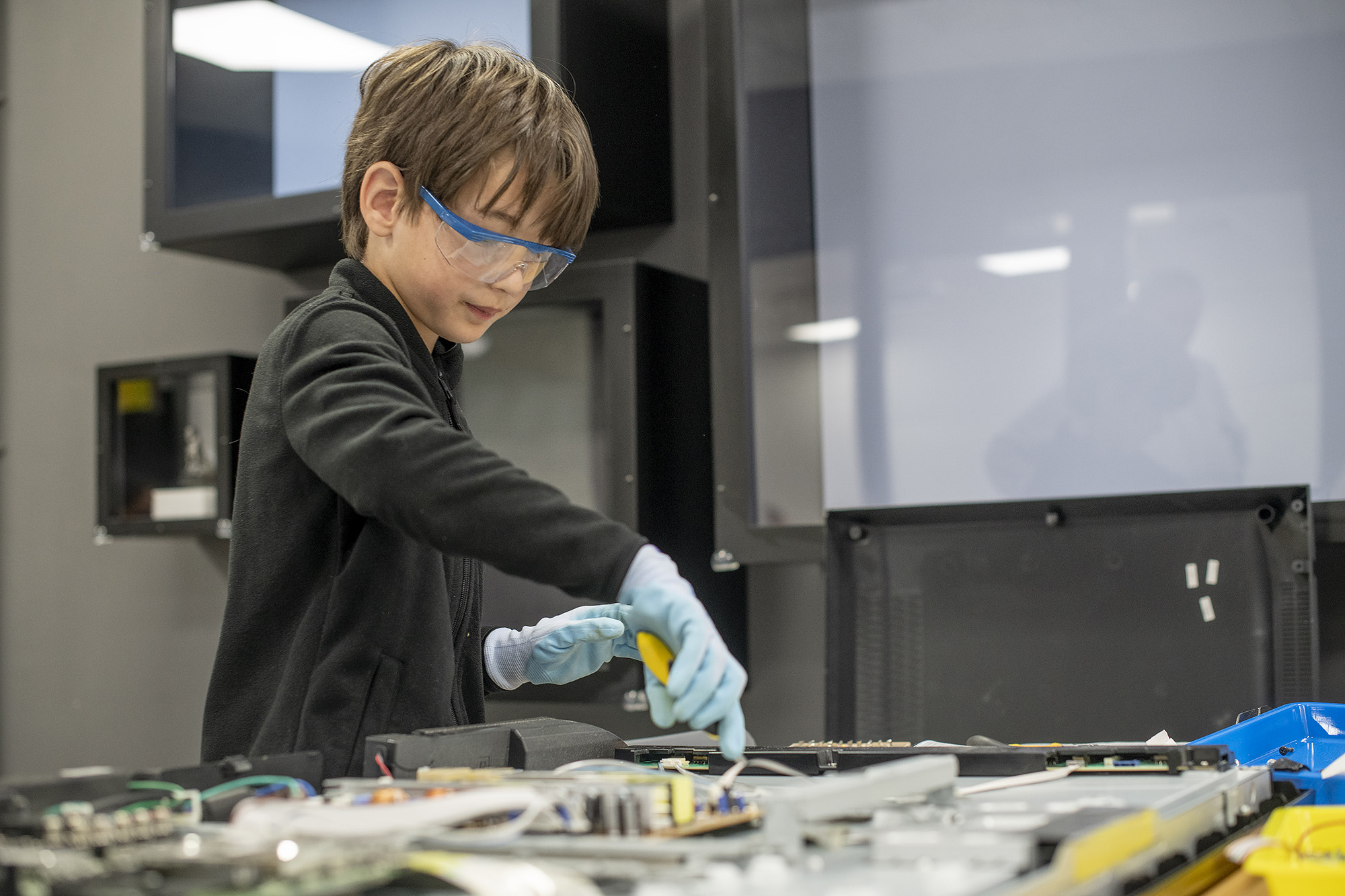 A boy wearing safety goggles and gloves is using tools to take apart an appliance on a worktop