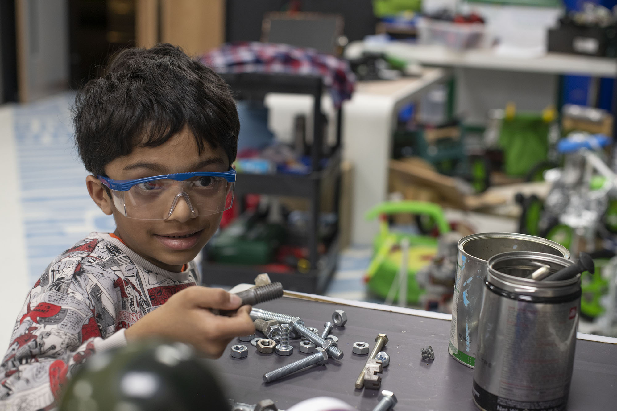 A boy wearing safety goggles is holding a metal bolt, picked up from a pile of nuts and bolts on the table in front of him.