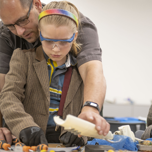 A man is helping a girl with tools and mechanical parts laid out on a table. The girl is wearing safety goggles, protective gloves and a rainbow hairband