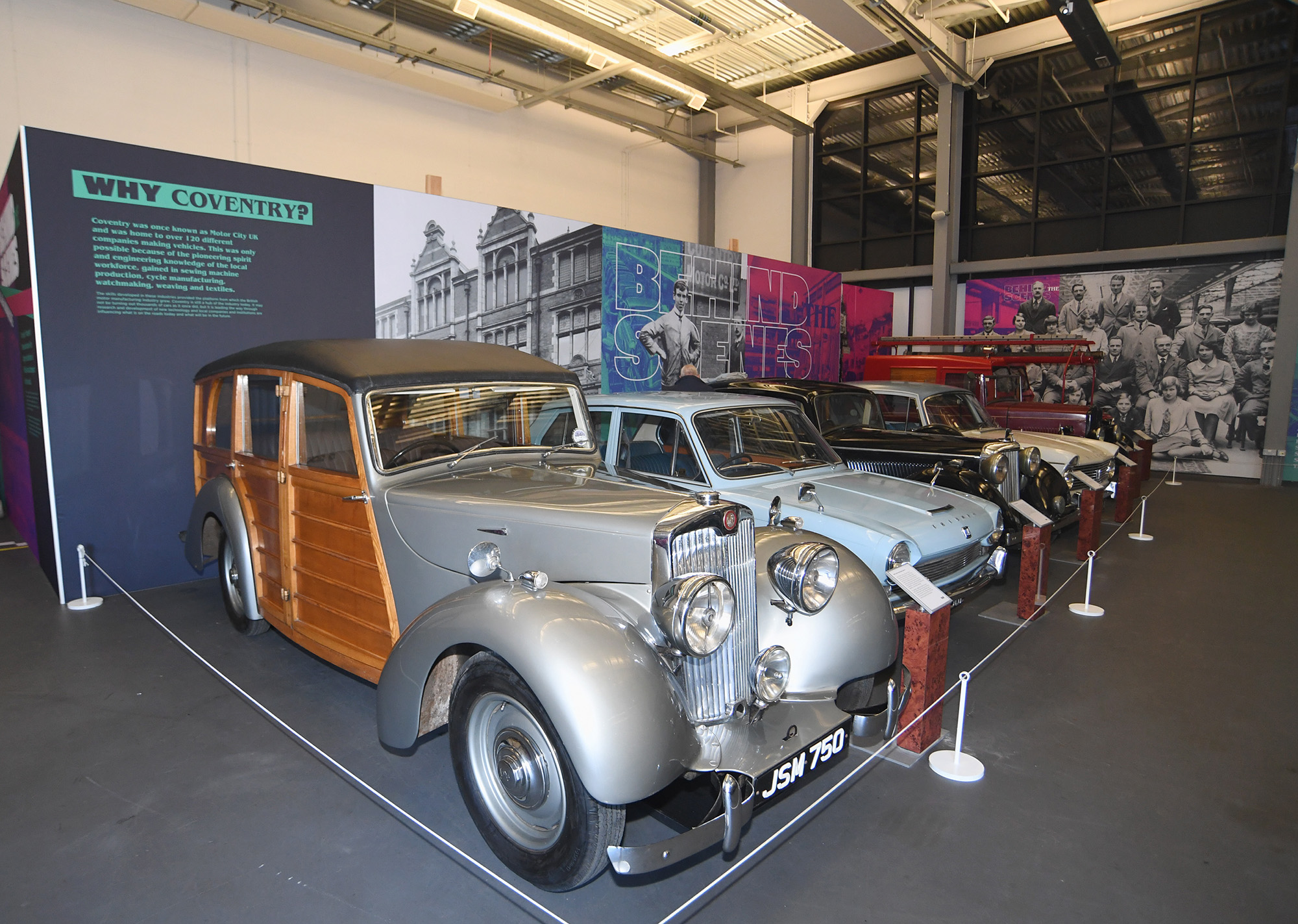 A selection of cars on display including a wooden-bodied Lea Francis estate in the foreground. A display board behind the cars features archive imagery of factory workers and the headlines, "Behind the Scenes" and "Why Coventry?"
