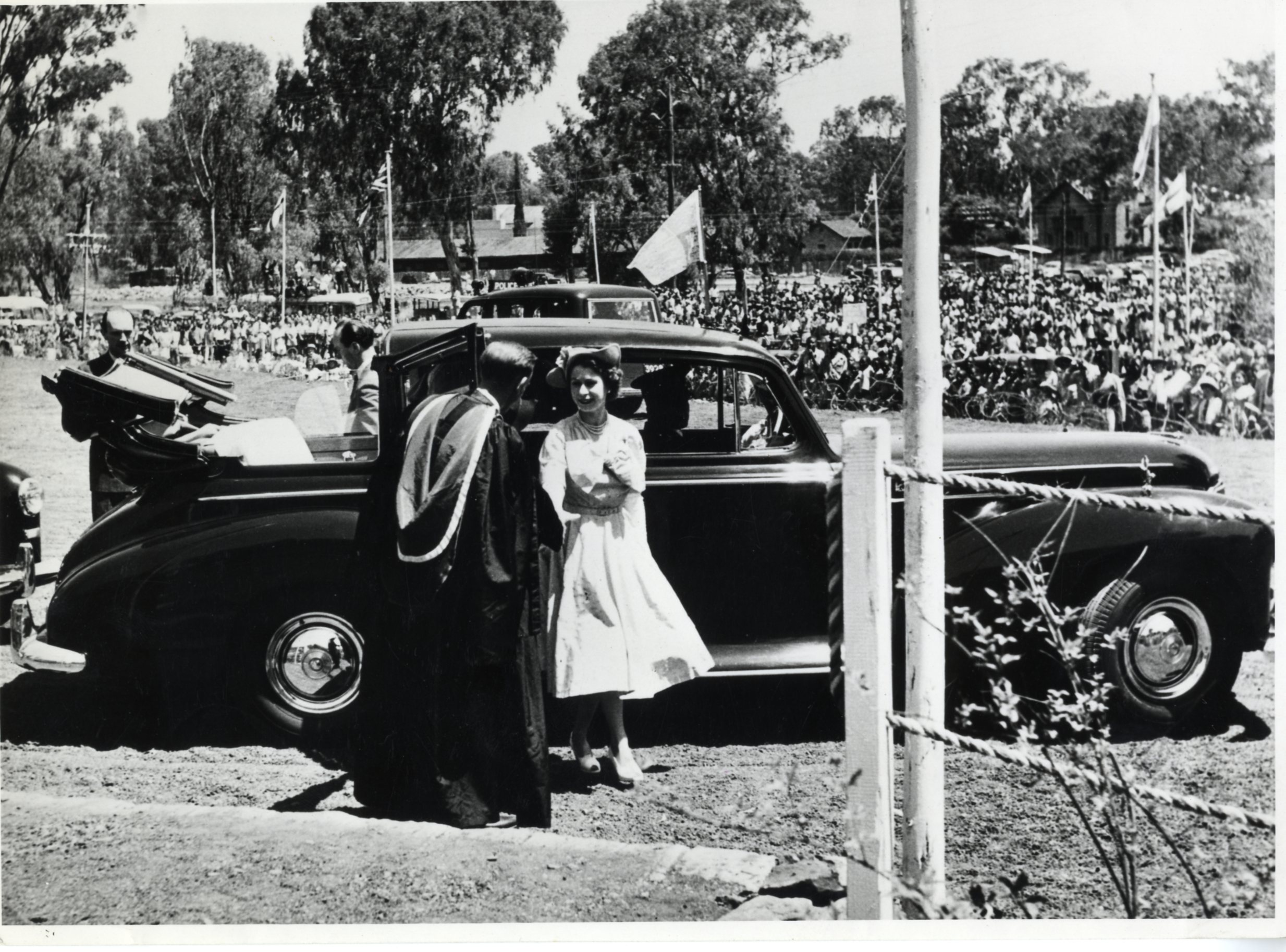 A black and white photograph of the queen stepping out of a car and being greeted by a man in ceremonial attire (possibly a priest). Crowds are gathered and flags are waving in the background behind the car.