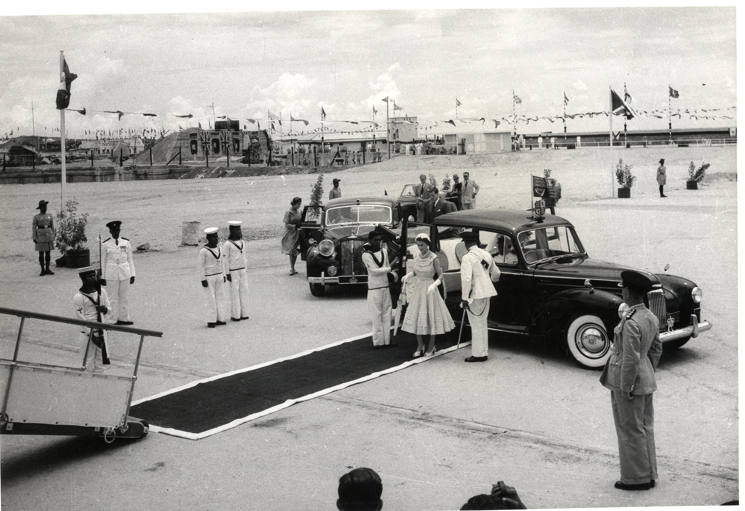 Queen Elizabeth II wearing a light summer dress, stepping out of the Humber Pullman Landaulette onto a carpet as a man holds open the door for her. Other men attend around the carpet and a ramp front left. Behind them is a large, flat expanse of ground. 