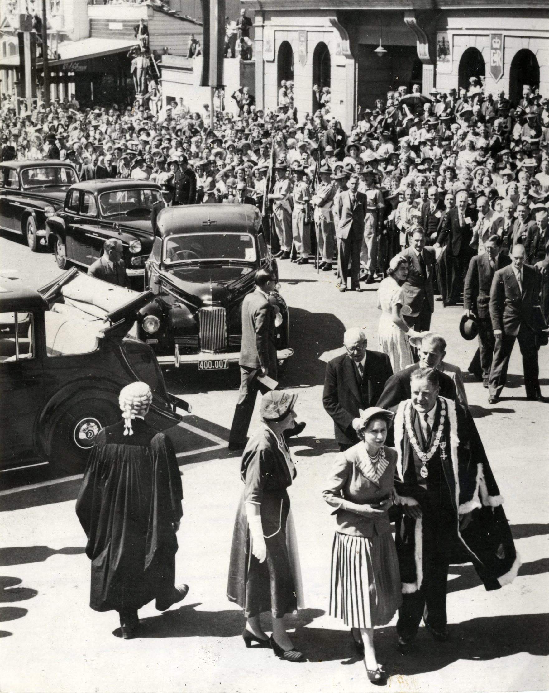 A black and white photograph of Queen Elizabeth II walking away from a long line of Humber cars, accompanied the Duke of Edinburgh and a small group of others including a man in a ceremonial gown and mayoral chain. Behind them, a large crowd of people has gathered beside the cars.