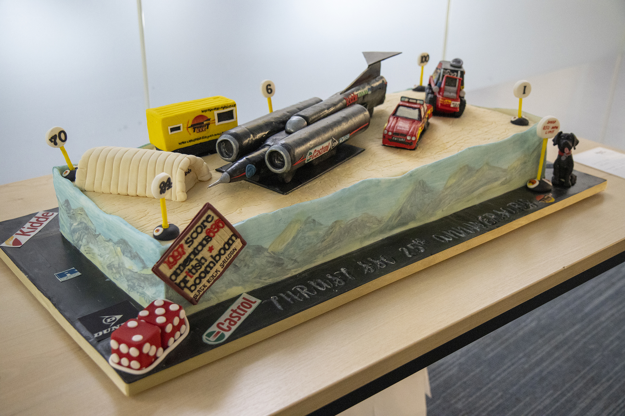 A huge cake with an icing model of Thrust SSC on the top. There are mountains painted on the side, as well as various other Thrust-related objects made out of icing, including support vehicles, mile markers, fluffy dice and a pet dog