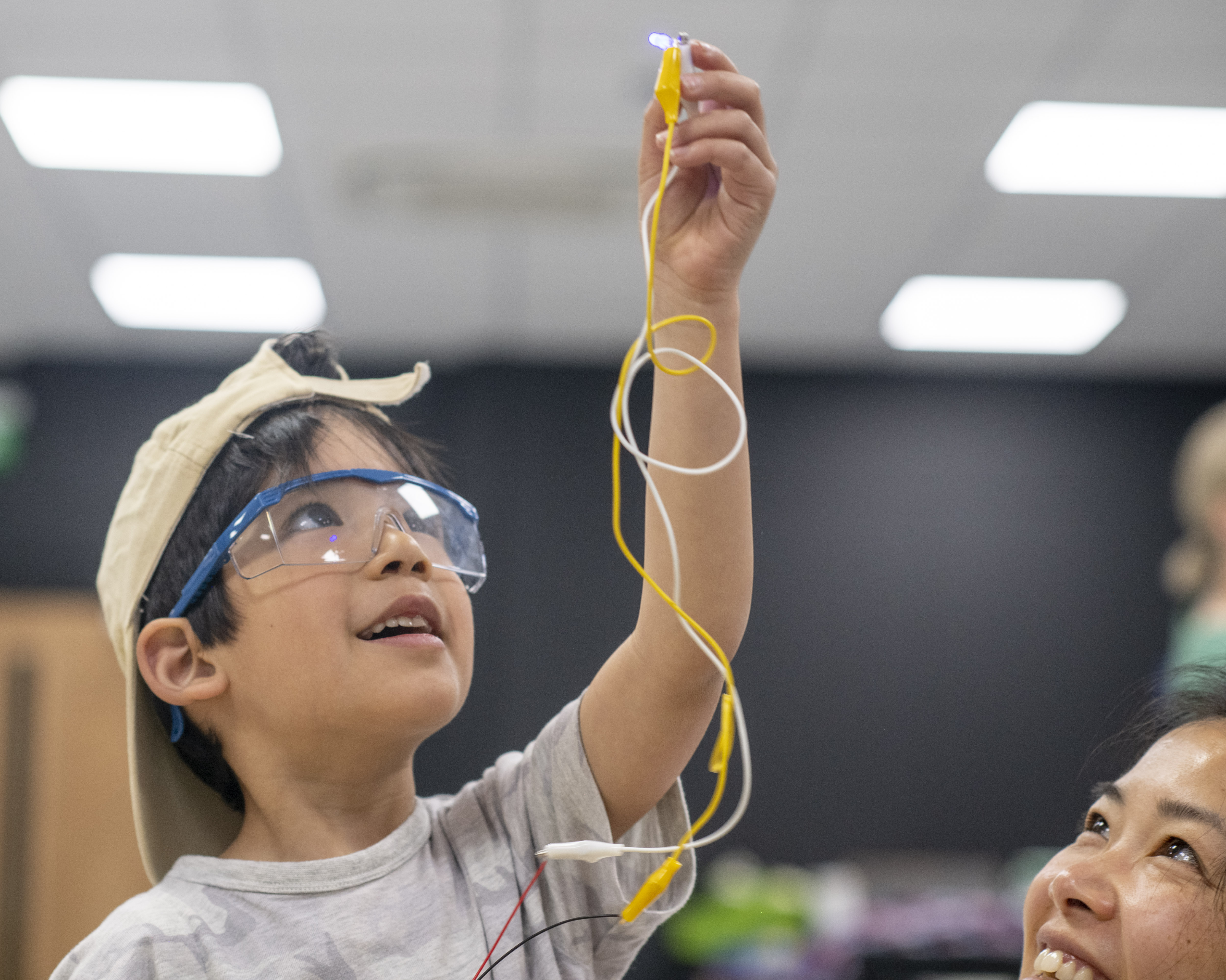 A boy wearing safety goggles and a backwards baseball cap holds up a pair of yellow and white cables with a crocodile clip at the end