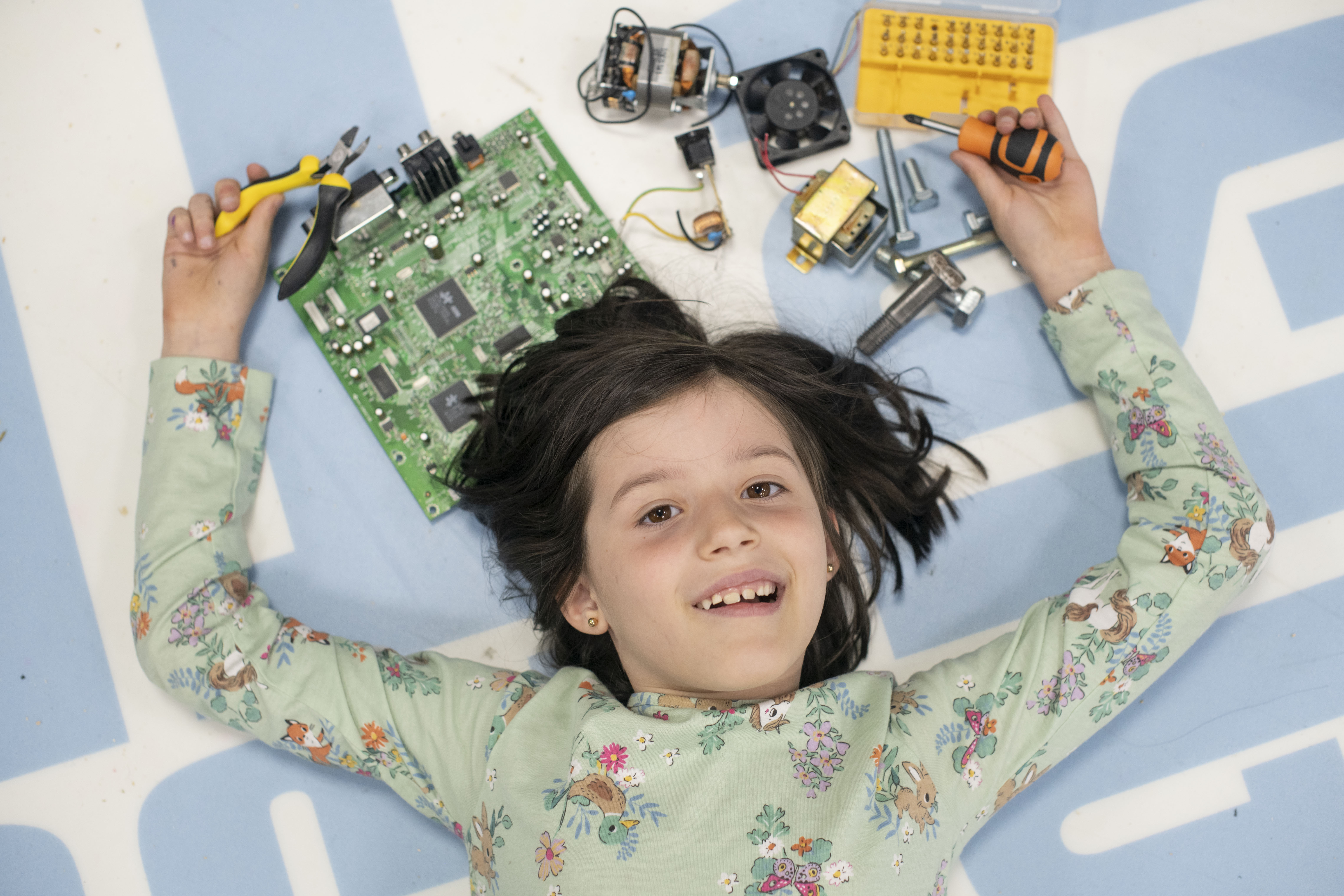 A girl lies on the floor surrounded by tools and electronic parts
